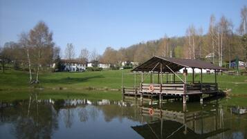 Une jetée en bois surmontée d’un abri, surplombant un lac paisible, entourée d’arbres et avec des maisons en arrière-plan.