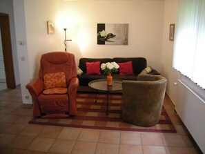 A living room with a brown leather armchair, a black sofa with red cushions, a round coffee table, and a large brown ottoman.