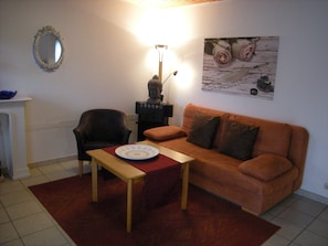 En stue med en sofa, en lænestol, et bord med en tallerken, en Buddha-statue, en lampe og et billede i ramme på væggen.