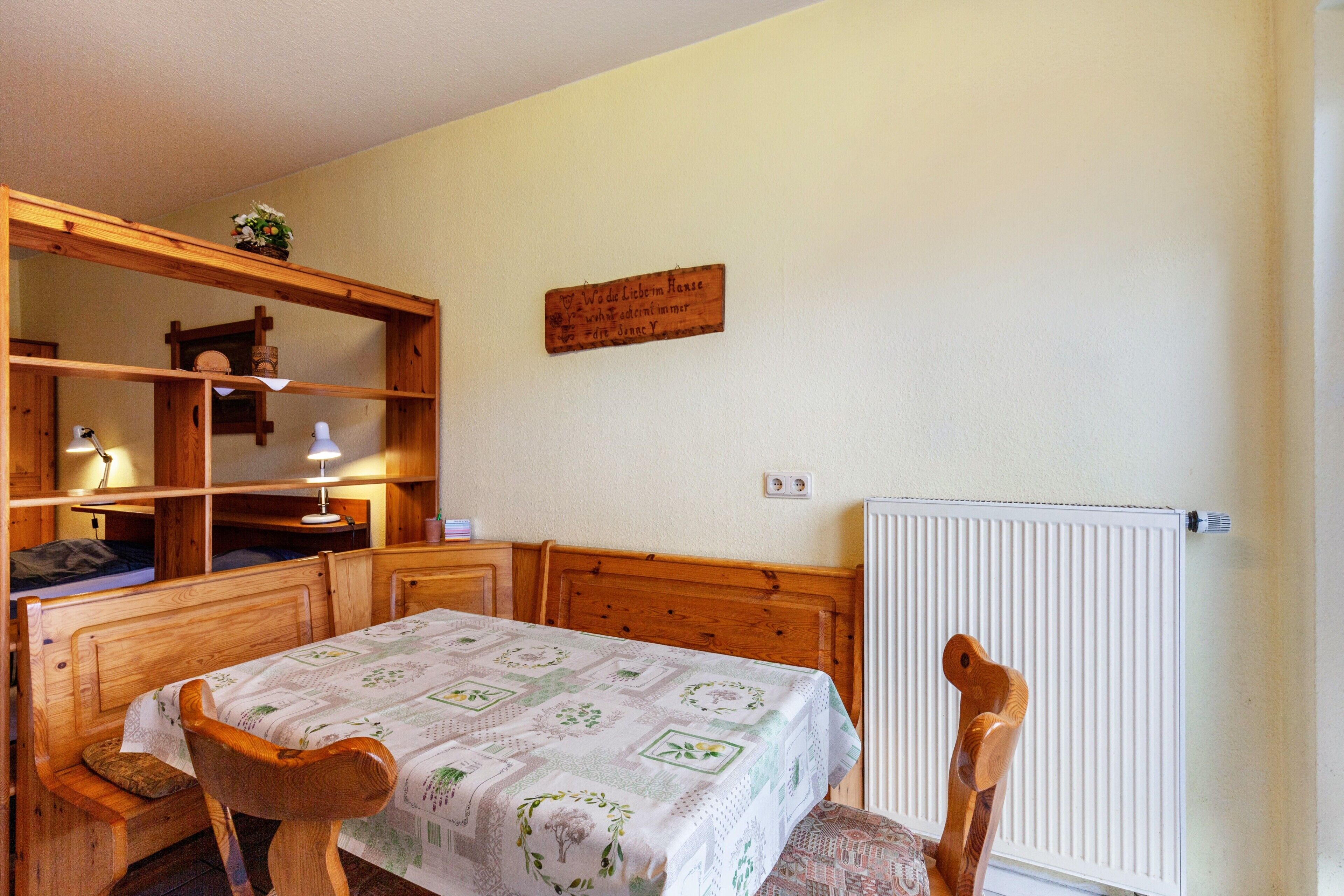A room with a wooden bed, a wooden desk, a chair, a radiator, and a shelf with a plant.