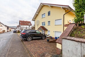 Une rue résidentielle bordée d’un SUV noir garé, d’une maison jaune avec un toit rouge et d’un chemin pavé en briques.