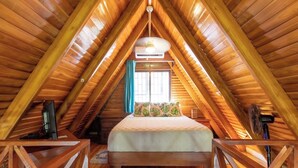 Une chambre mansardée chaleureuse avec un plafond en bois, un lit recouvert d’une courtepointe à motifs floraux et une fenêtre agrémentée de rideaux.