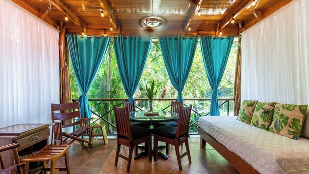 Une pièce avec un plafond en bois, un lit agrémenté d’oreillers à motifs de feuilles vertes, une table ronde avec des chaises, et une vue sur la verdure à travers des rideaux bleus.