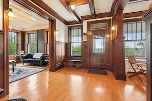 A spacious room with wooden floors, a black leather sofa, a wooden chair, and a large window with a view of greenery.