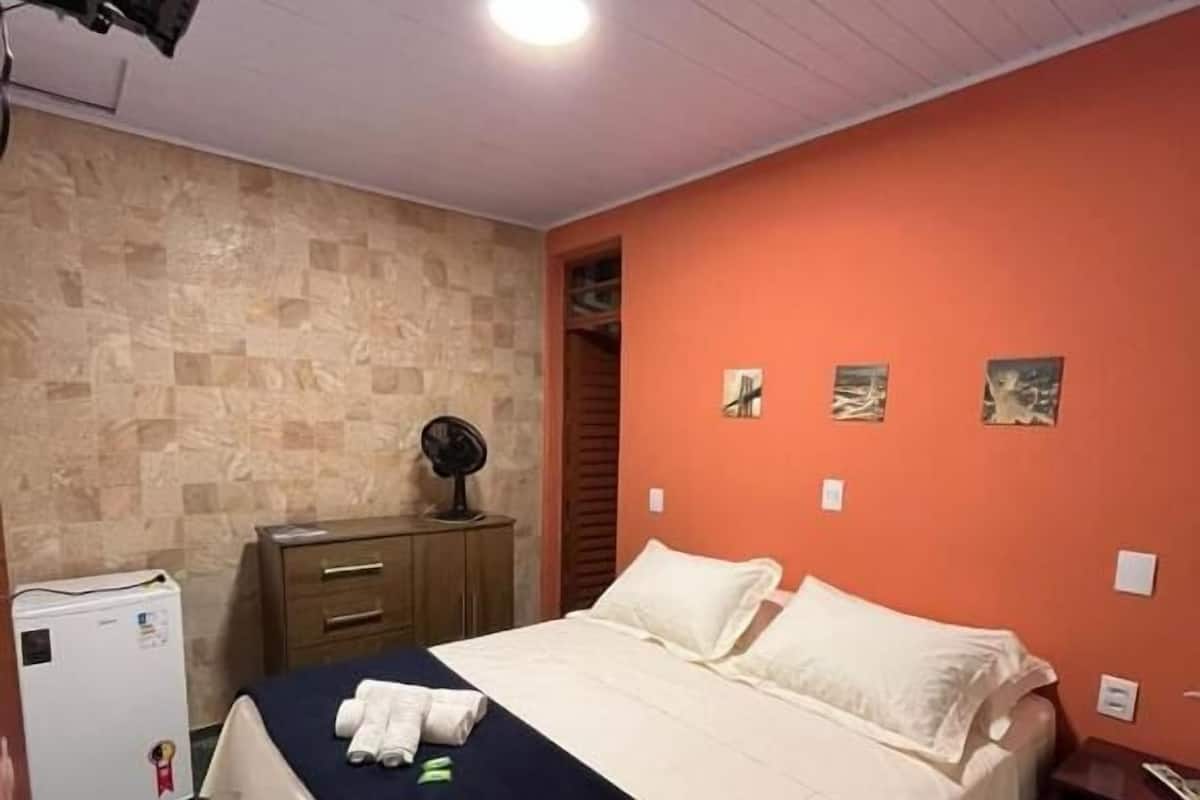 A hotel room with a bed, a small desk, a fan, and a refrigerator.