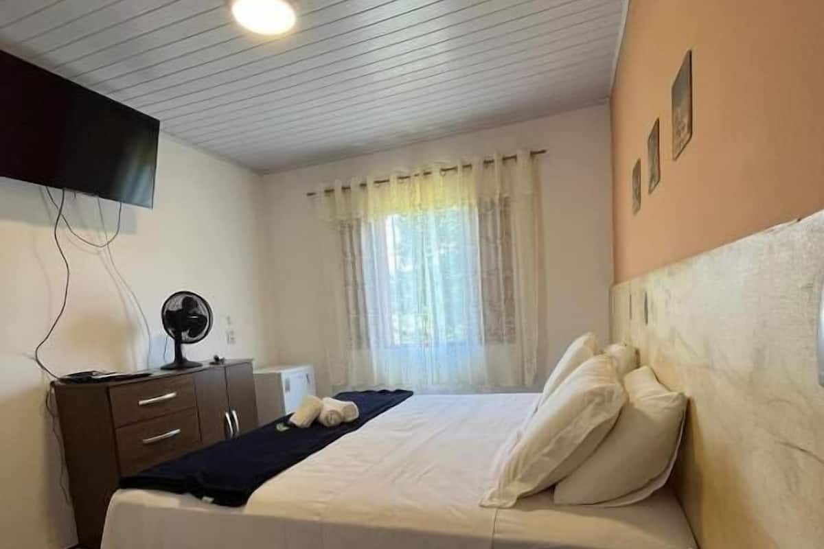 A hotel room with a bed, a desk, a chair, a flat-screen TV mounted on the wall, a window with curtains, and a ceiling light.