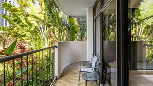 Un balcone con pavimento piastrellato, un tavolo in vetro e sedie, circondato da rigogliosa vegetazione tropicale e ringhiera metallica.