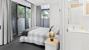 Una camera d'albergo moderna con un letto grande, un comodino con una lampada, un bagno bianco con uno specchio e una porta scorrevole in vetro che dà su uno spazio all'aperto.