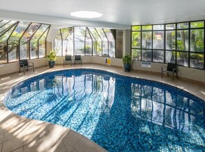 An indoor swimming pool with clear blue water, surrounded by large windows offering views of greenery. - Alexander Apts - Ground Floor 2 Bedroom (Surfers Paradise)