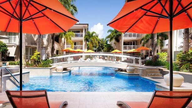 A pool area with lounge chairs and umbrellas, a bridge, and a building with balconies.