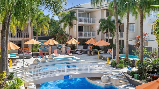 A pool area with lounge chairs and umbrellas, surrounded by palm trees and a multi-story building.