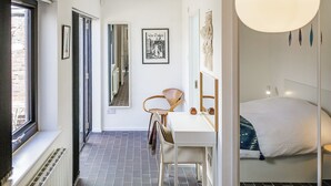 A modern hotel room with a large bed, a desk with a chair, a window, and a door leading to another room. - Mid-Century Modern Style 1BD with Garden (Cheltenham)