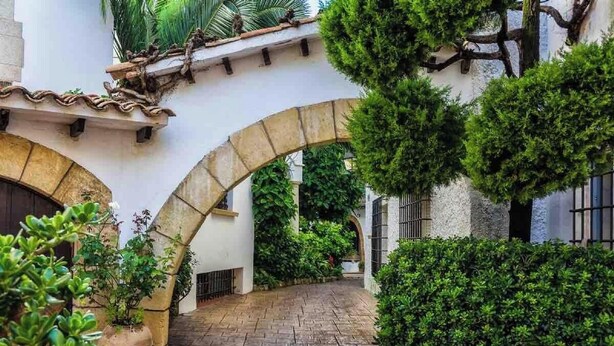 A courtyard with a stone archway, lush greenery, and a tiled roof. - Holiday Home 'Chalet Independiente Costa Daurada' with Private Garden, Balcony and Air Conditioning (Roda de Berà)