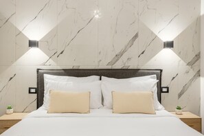 Une chambre d’hôtel avec un mur décoré d’un motif imitant le marbre, un lit avec une literie blanche et deux oreillers beige, et deux lampes fixées au mur.