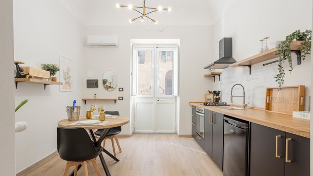 Una cocina moderna con comedor, armarios de madera, lavavajillas negro y una planta colgante.