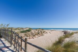 En træpromenade der fører til en sandstrand med klitter og havet i baggrunden.