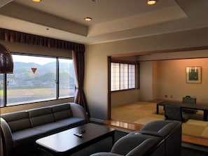 Ein modernes Hotelzimmer mit einer Couch, einem kleinen Tisch, einem Esstisch mit Stühlen, einem großen Fenster mit Blick auf die Landschaft und einer traditionellen Schiebetür.