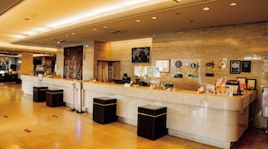 Zona de recepción de hotel con mostrador de mármol, una recepcionista y varios paneles informativos.