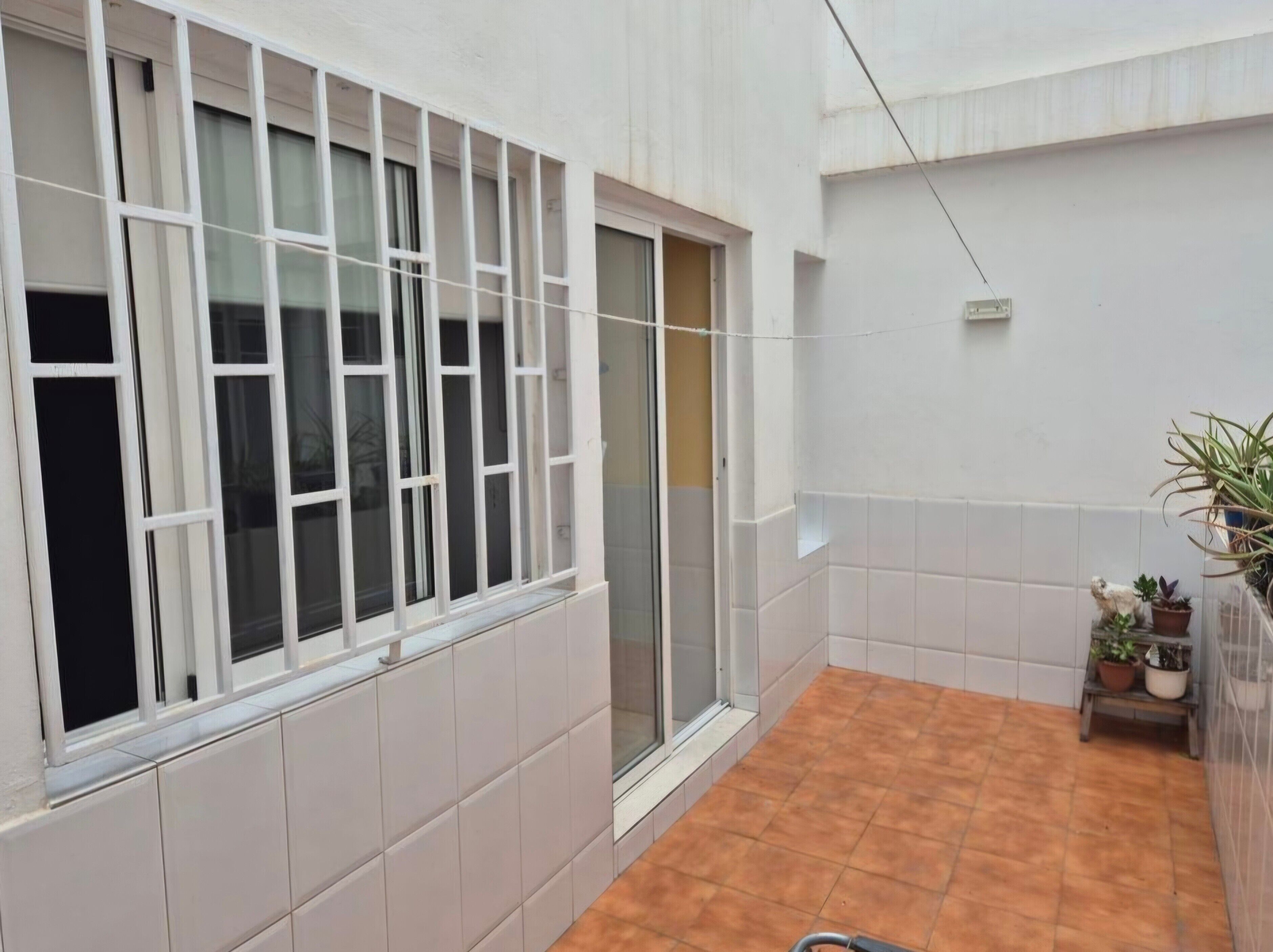 A room with white tiled walls, a window with white frames, a sliding door, and a tiled floor. There are potted plants on a small table.