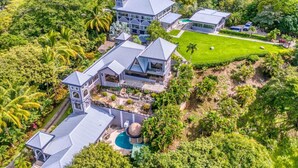 Une grande maison moderne, dotée de plusieurs annexes, d’une piscine et entourée d’une végétation luxuriante.