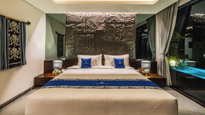 A modern hotel room with a large bed, a decorative headboard, a nightstand with a lamp, and a view of a water feature. - De Prestigio Villa I (Canggu)