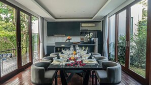 A dining area with a table set for six, surrounded by chairs, and large windows offering a view of greenery. - De Prestigio Villa I (Canggu)