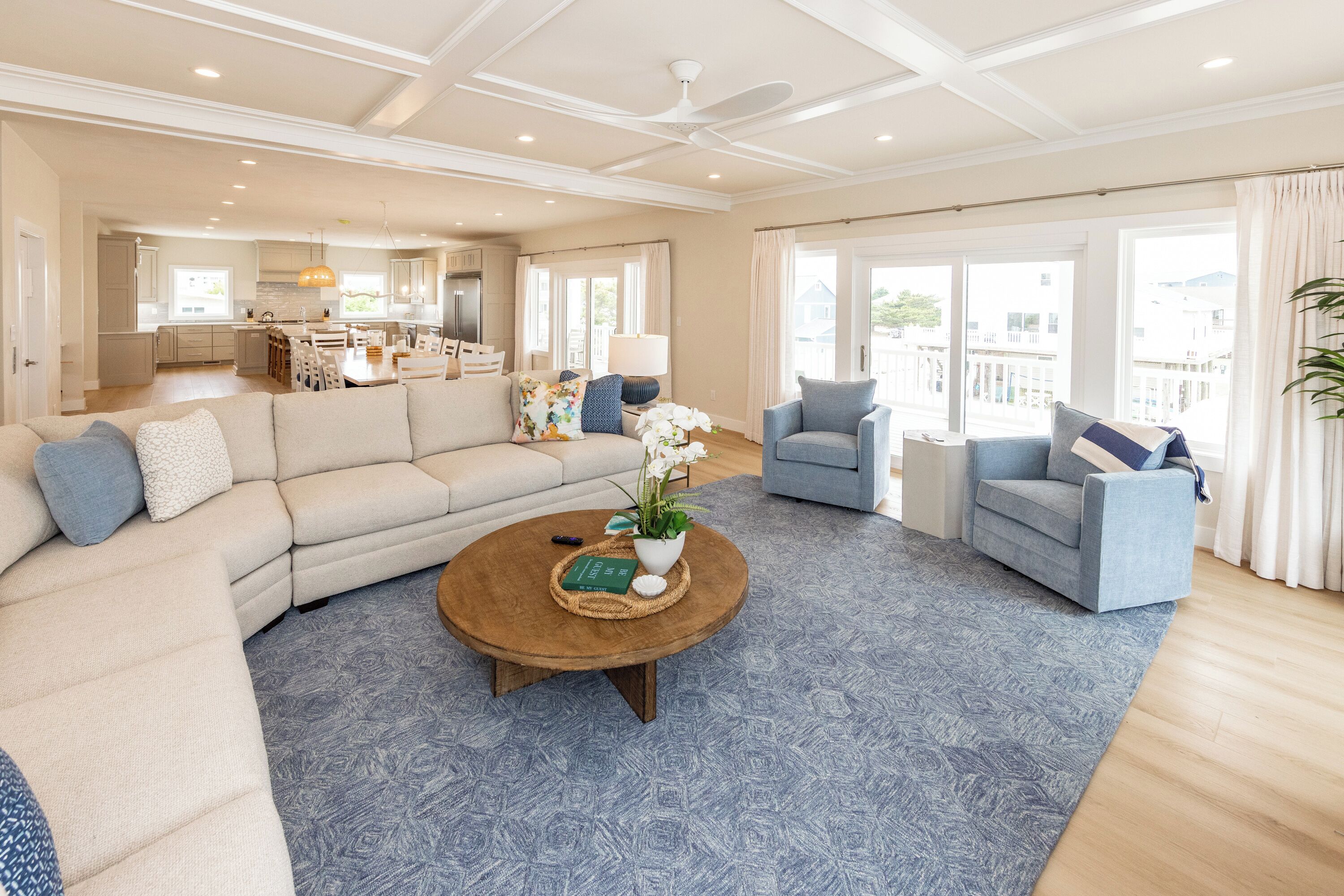A spacious living room with a large sectional sofa, a round coffee table with decorative items, and a view of the kitchen and balcony.