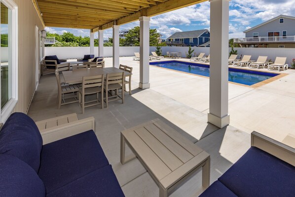 A covered outdoor area with a pool, lounge chairs, and a wooden table and chairs.