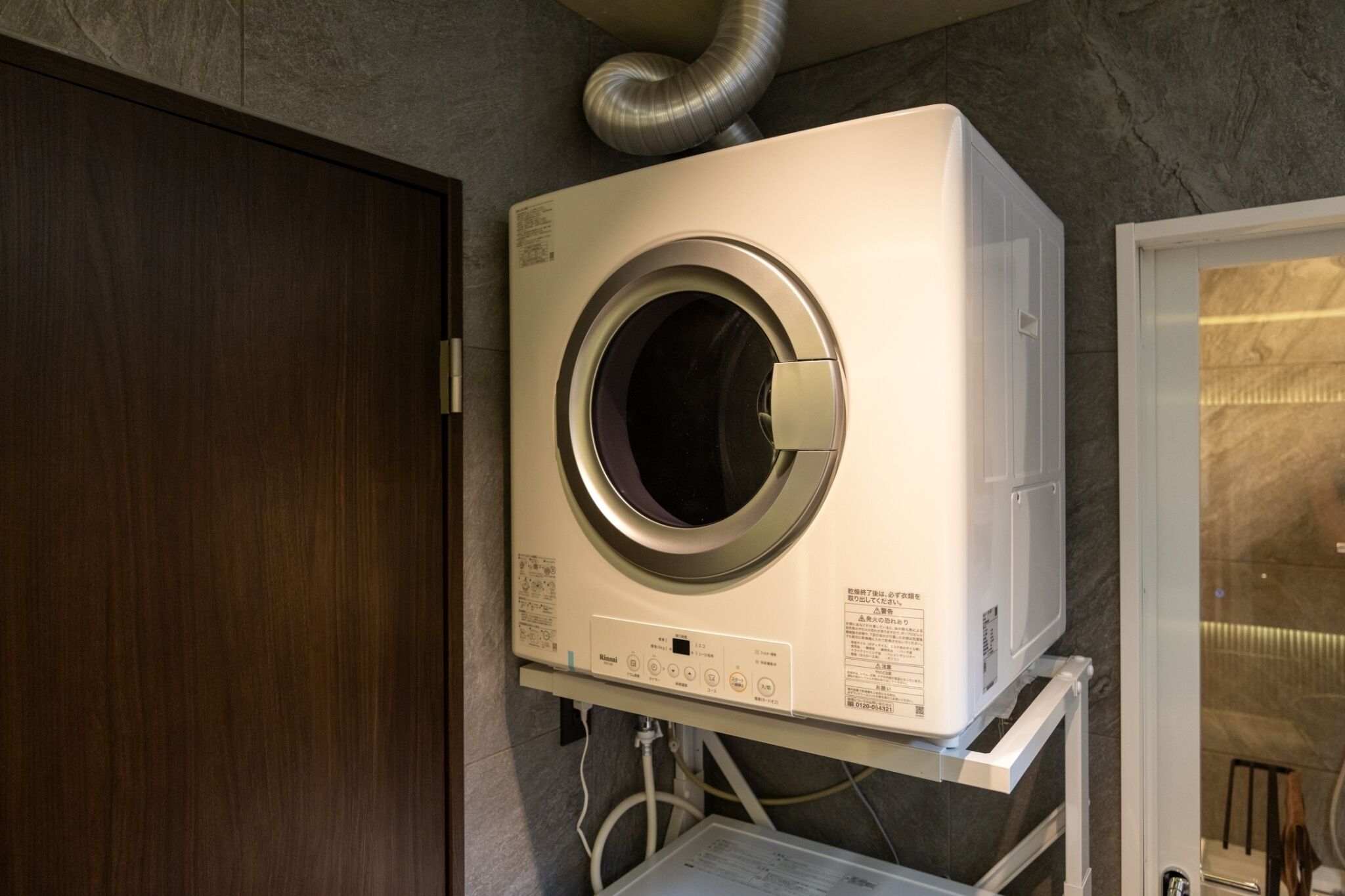 A compact, wall-mounted washer and dryer unit in a bathroom with a wooden door and a glass shower enclosure.