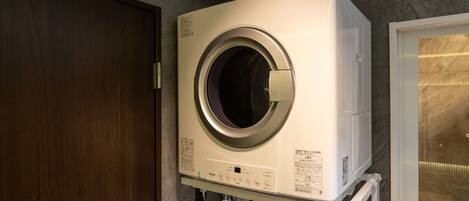 A compact, wall-mounted washer and dryer unit in a bathroom with a wooden door and a glass shower enclosure.