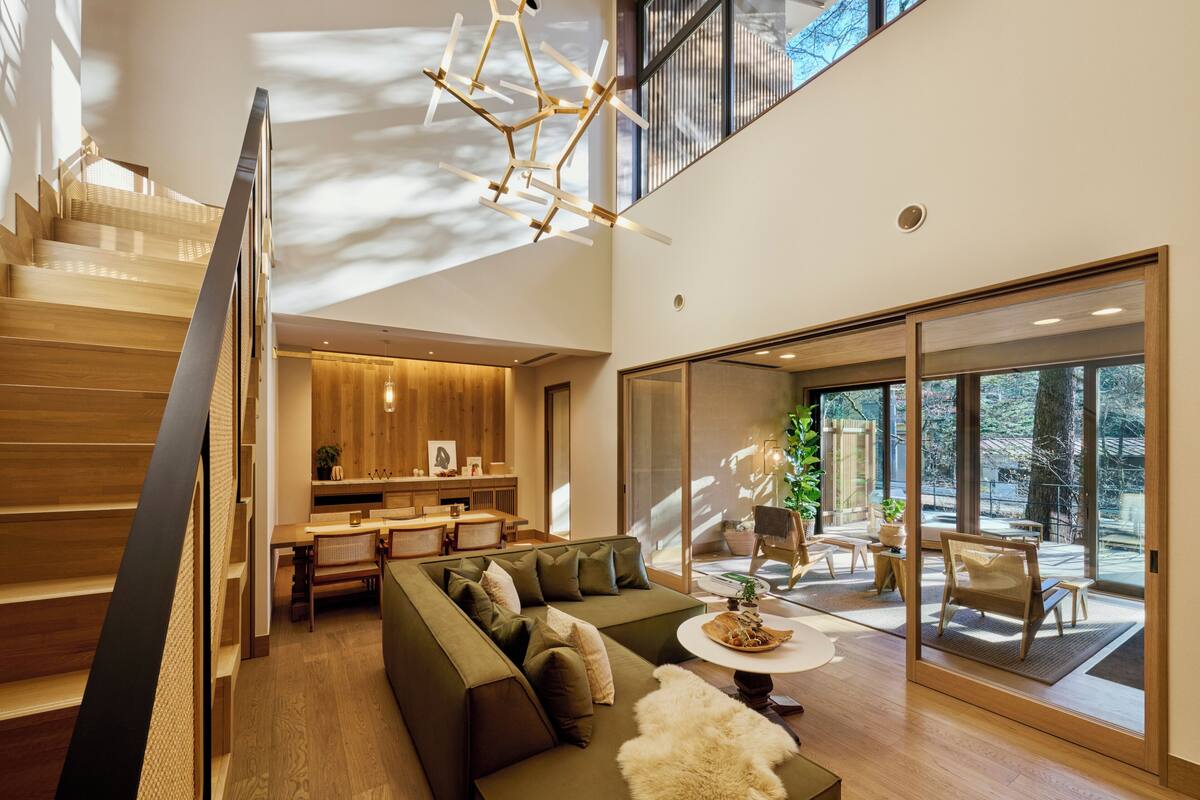 A modern interior with a staircase, a green sofa, a dining area, and large windows offering a view of the outdoors.