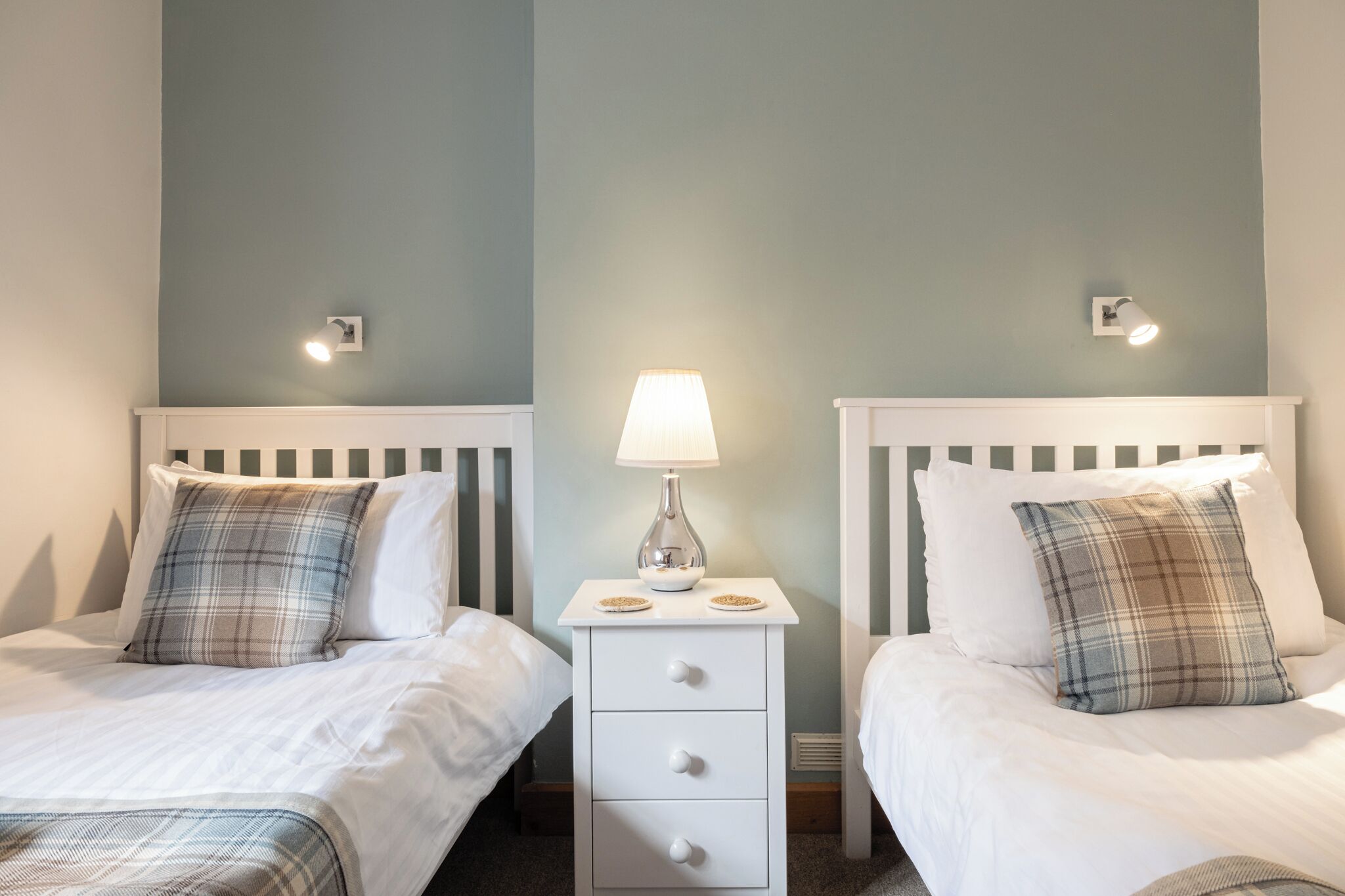 Habitación de hotel con dos camas, cada una con ropa blanca y almohadas de cuadros. Se ve una mesita de noche con una lámpara y un frutero.
