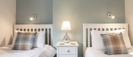 Habitación de hotel con dos camas, cada una con ropa blanca y almohadas de cuadros. Se ve una mesita de noche con una lámpara y un frutero.