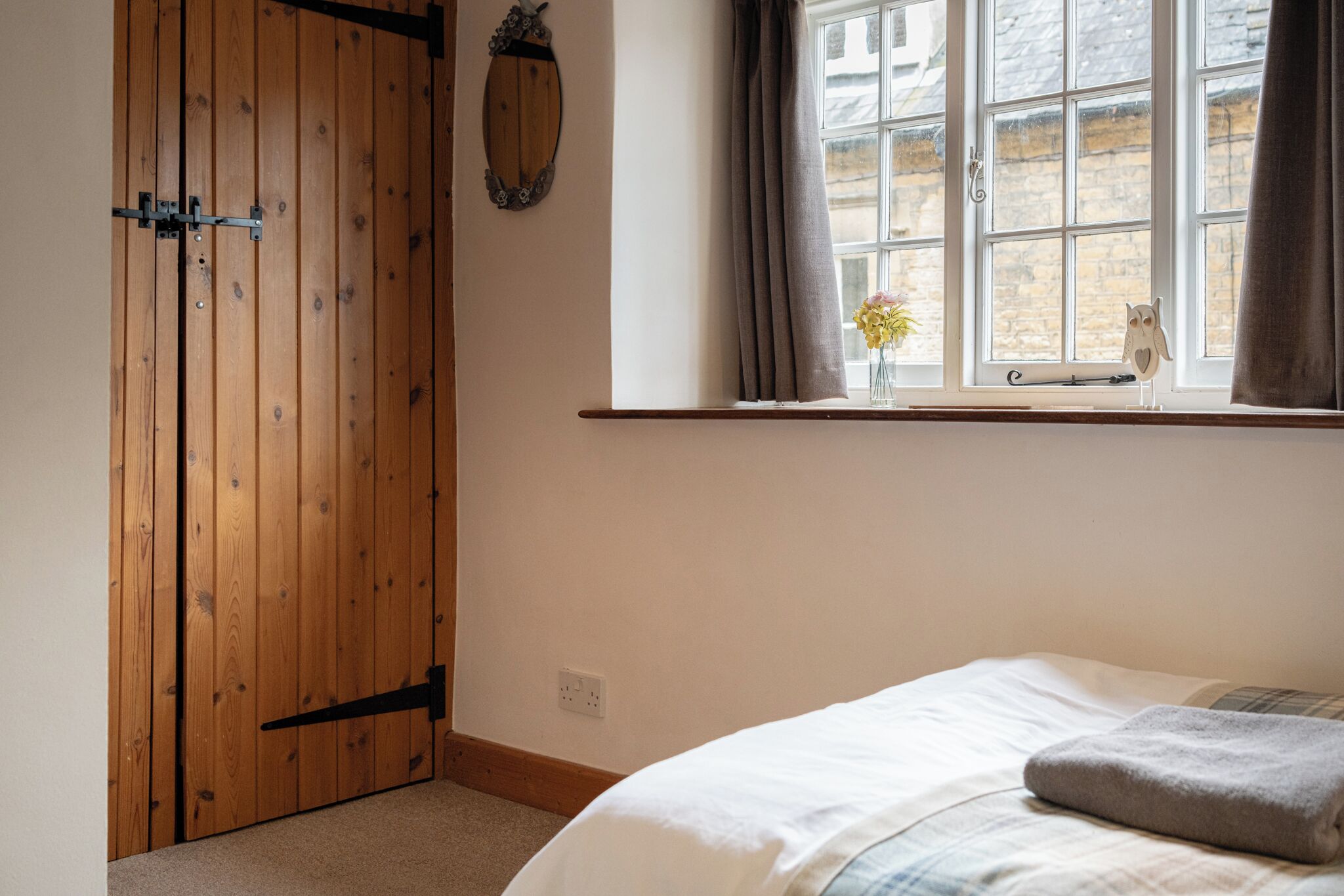 Un dormitorio con un armario de madera, una ventana con cortinas, una mesita de noche con un florero y una cama con un almohadón.