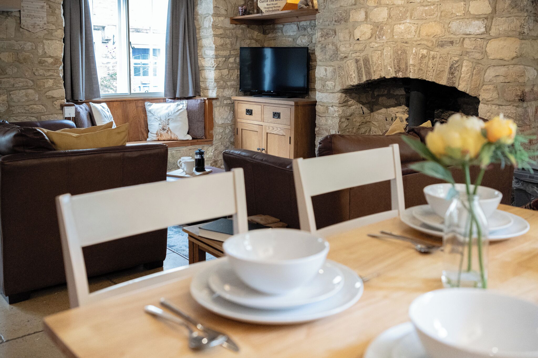 Un salon chaleureux avec un mur en pierre, une cheminée, un téléviseur et un coin repas où une table est dressée pour deux personnes.