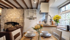 A cozy kitchen with a stone fireplace, wooden beams, white cabinets, and a window with flowers. - Maple Cottage - Beautiful 2BD Retreat (Chipping Norton)