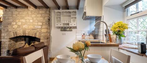 A cozy kitchen with a stone fireplace, wooden beams, white cabinets, and a window with flowers.