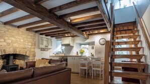 A cozy living room with a fireplace, a staircase, and a kitchen area. - Maple Cottage - Beautiful 2BD Retreat (Chipping Norton)