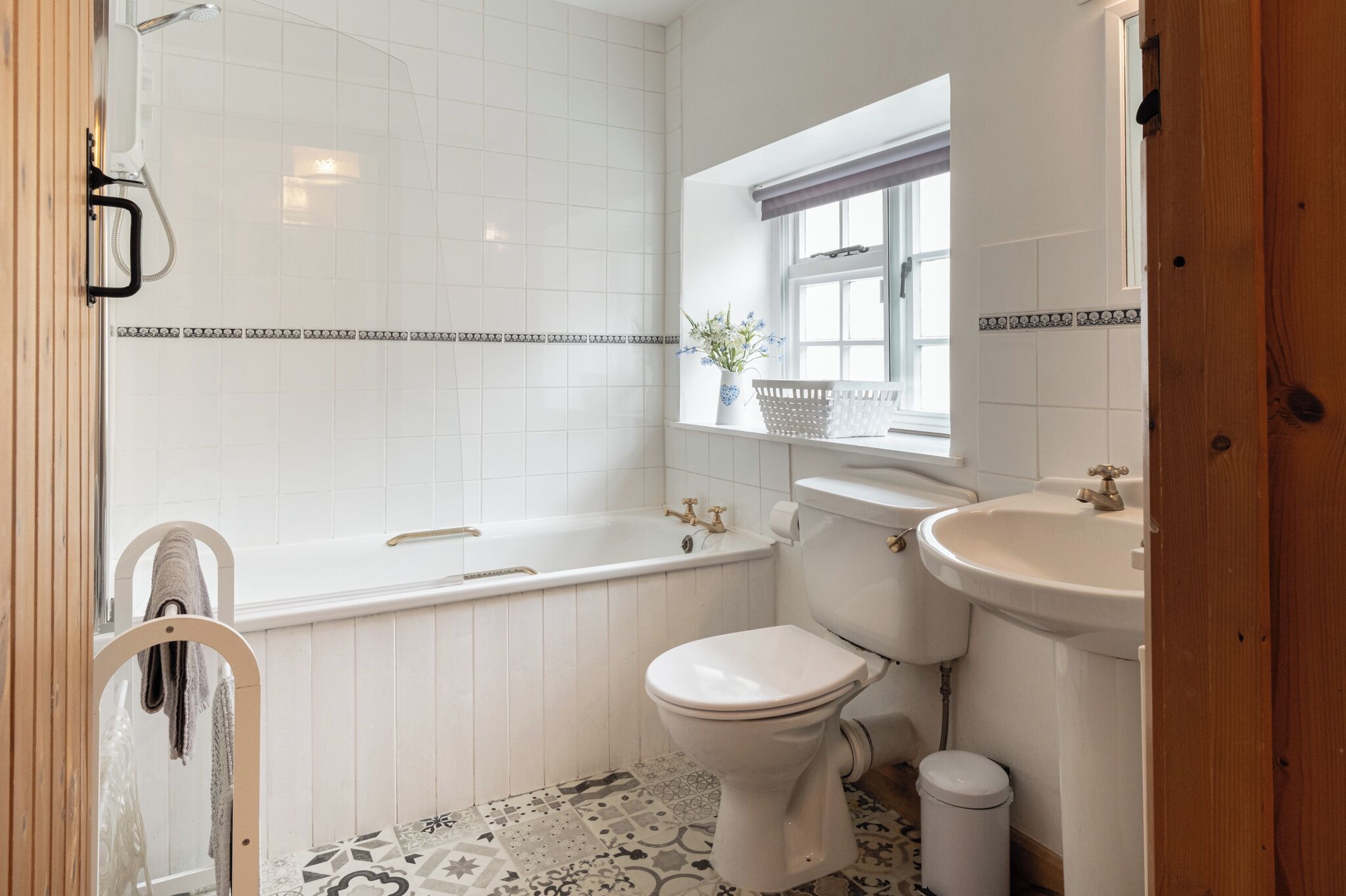 Et badeværelse med badekar, toilet og håndvask, med et mønstret gulv og et vindue med en plante.