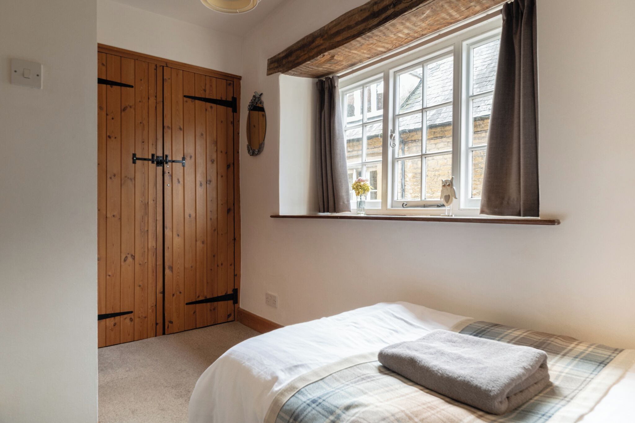 A bedroom with a bed, a window with curtains, a wooden wardrobe, and a small shelf with a vase.