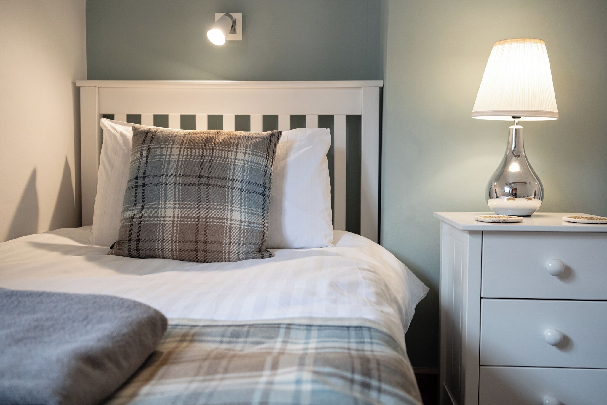 A neatly made bed with plaid and white pillows, a bedside lamp, and a white nightstand with drawers.