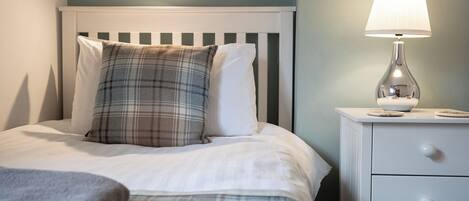 A neatly made bed with plaid and white pillows, a bedside lamp, and a white nightstand with drawers.