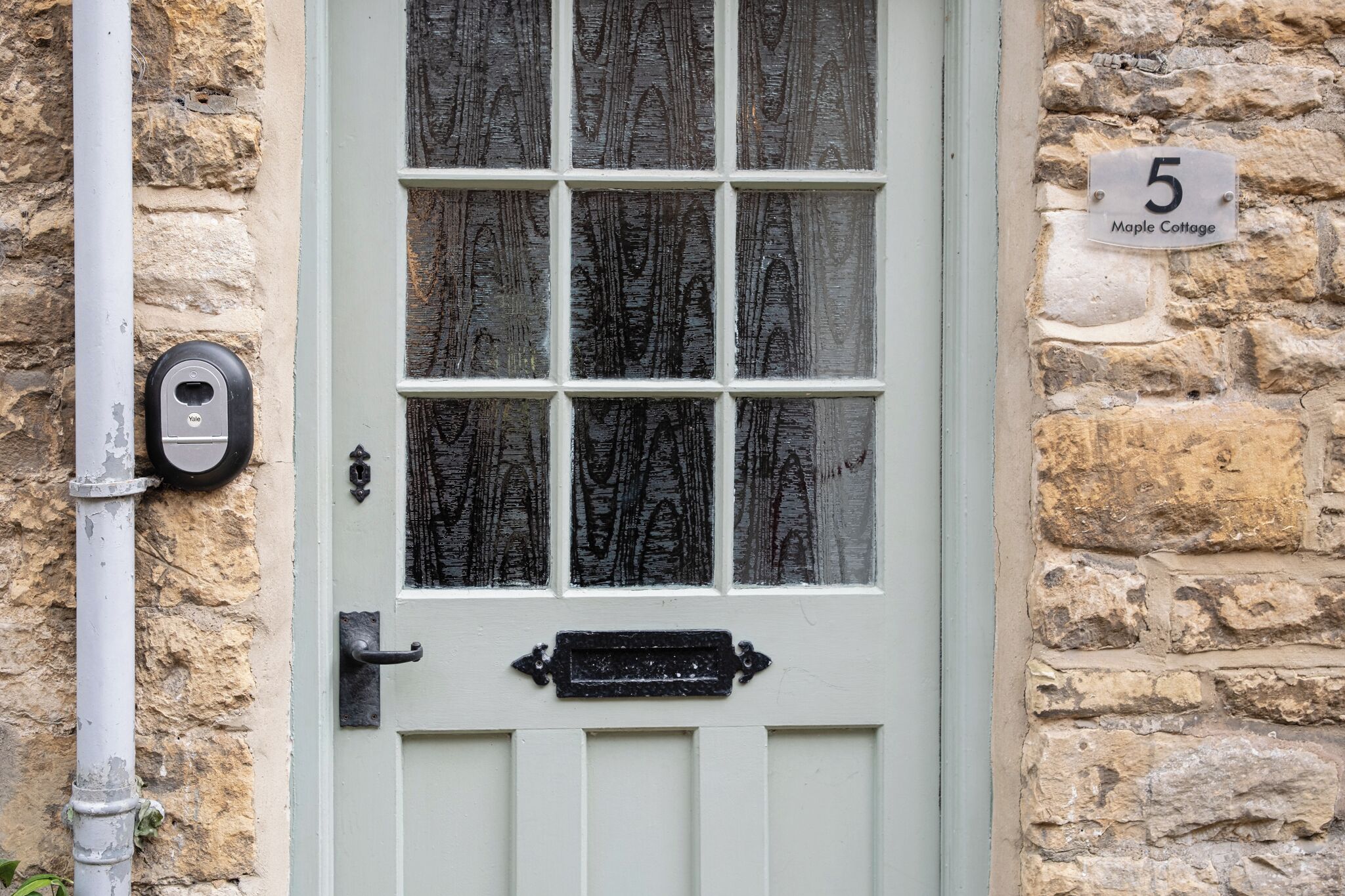 Una puerta blanca con manija negra y una ventana con vidrio esmerilado. Se ve una placa con el número "5 Maple Cottage" en la pared a la derecha.