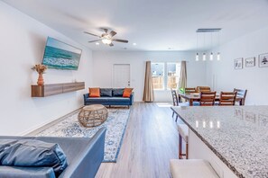 Una sala moderna con un sofá azul, una zona de comedor con sillas de madera, una cocina con encimera de granito y un ventilador de techo.