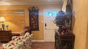 Una habitación con piso de ladrillo, un sofá floral, una mesita de madera con una lámpara y una puerta con ventana.