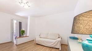 Una habitación minimalista con piso de madera, un sofá tapizado, un lámpara en el techo y una pequeña planta cerca de la puerta.