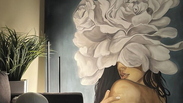 A room with a large floral mural on the wall, a potted plant, a sofa, and a small table with a lamp.