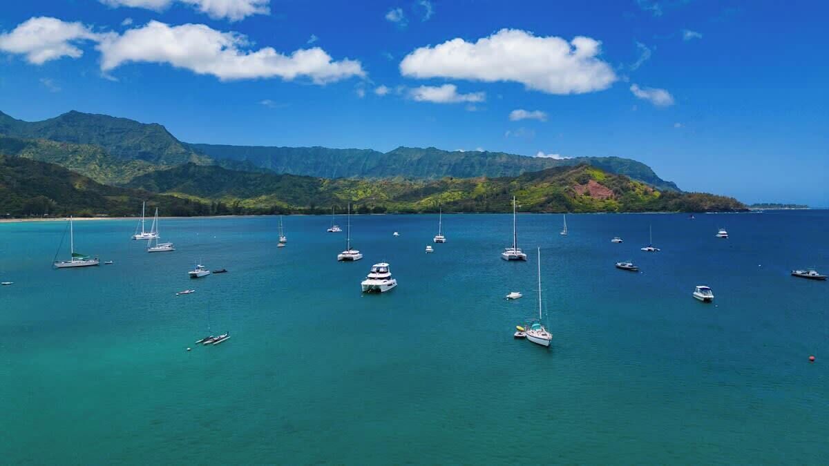 A coastal area with multiple boats anchored in clear turquoise waters, surrounded by lush green mountains under a blue sky with scattered clouds.