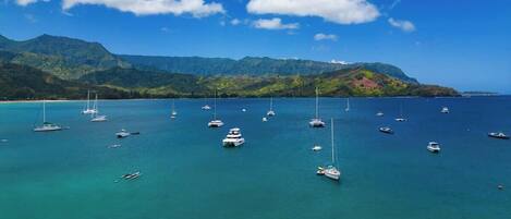 A coastal area with multiple boats anchored in clear turquoise waters, surrounded by lush green mountains under a blue sky with scattered clouds.
