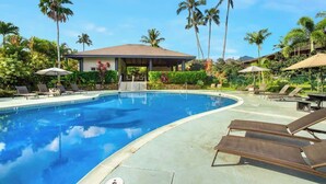 Ein Swimmingpool mit Liegestühlen und Sonnenschirmen, umgeben von Palmen und einem überdachten Pavillon.